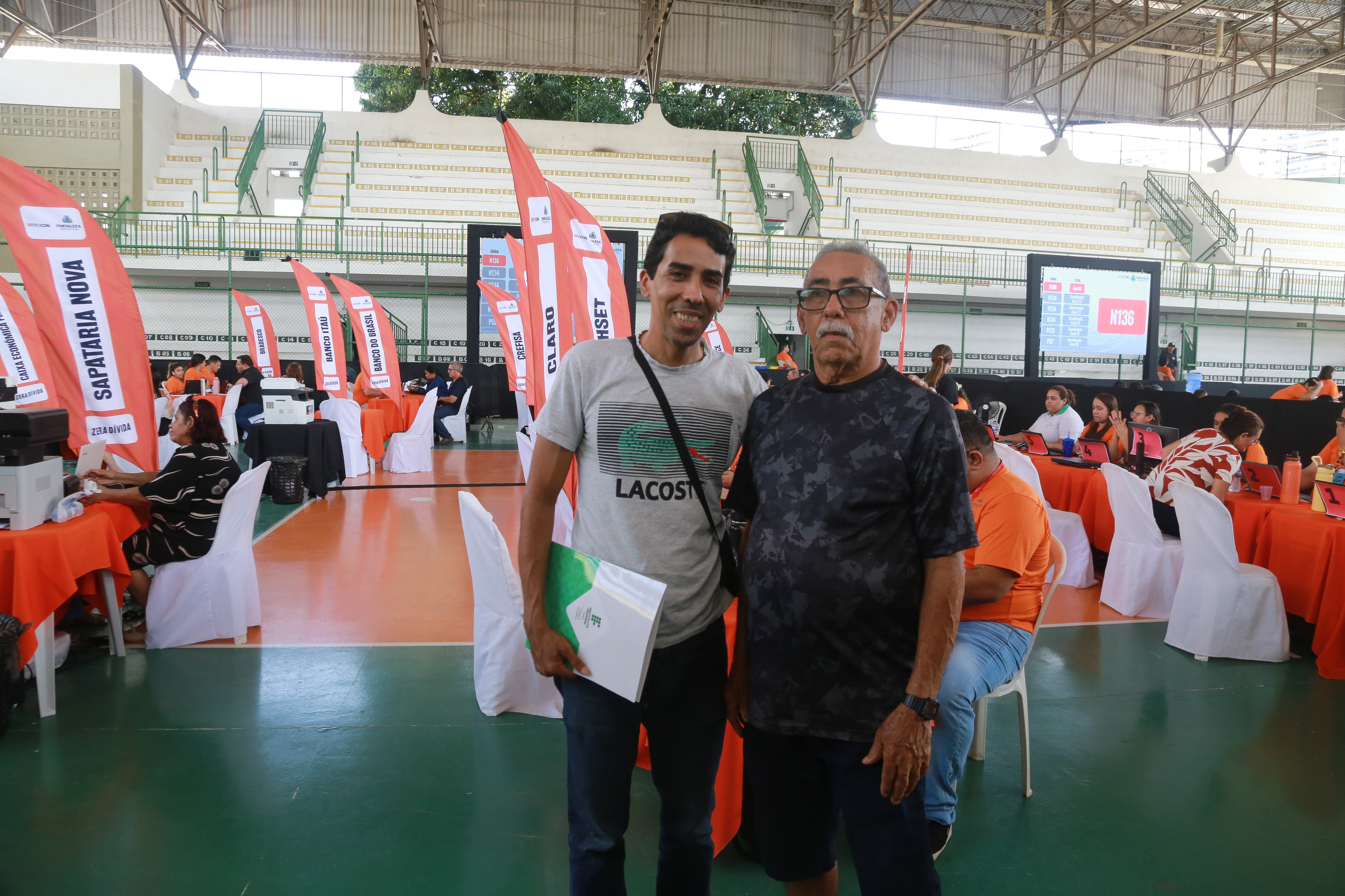 dois homens posam para a foto na quadra do ginásio aécio de borba; ao fundo as mesas de atendimento do Procon